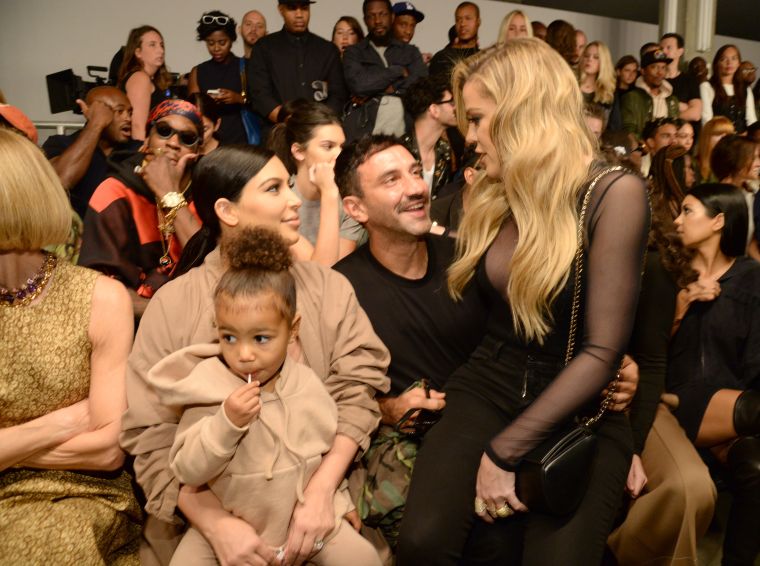 North West sucks on her lollipop while Kim talks to Khloe, sitting on Riccardo Tisci’s lap.