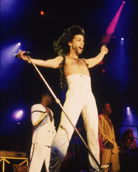 American singer and songwriter Prince stands on stage with his arms outstretched, wearing high-waisted white pants, circa 1990.