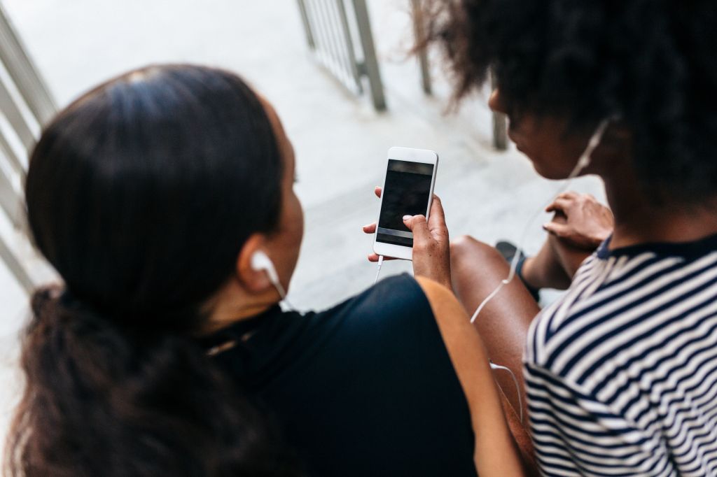 Back view of two friends sitting on stairs listening music together with earphones and smartphone