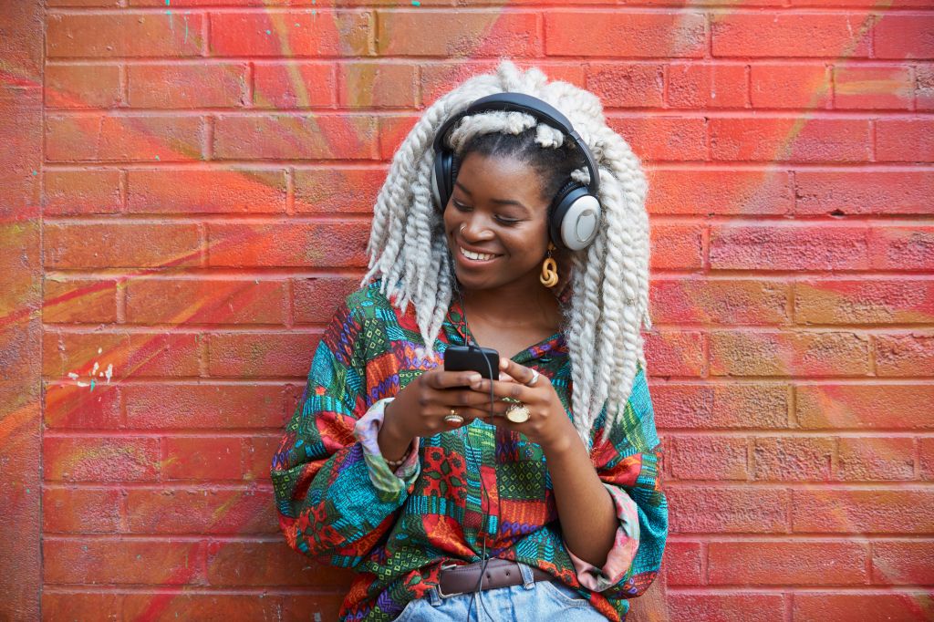 Black woman leaning on brick wall listening to cell phone with headphones