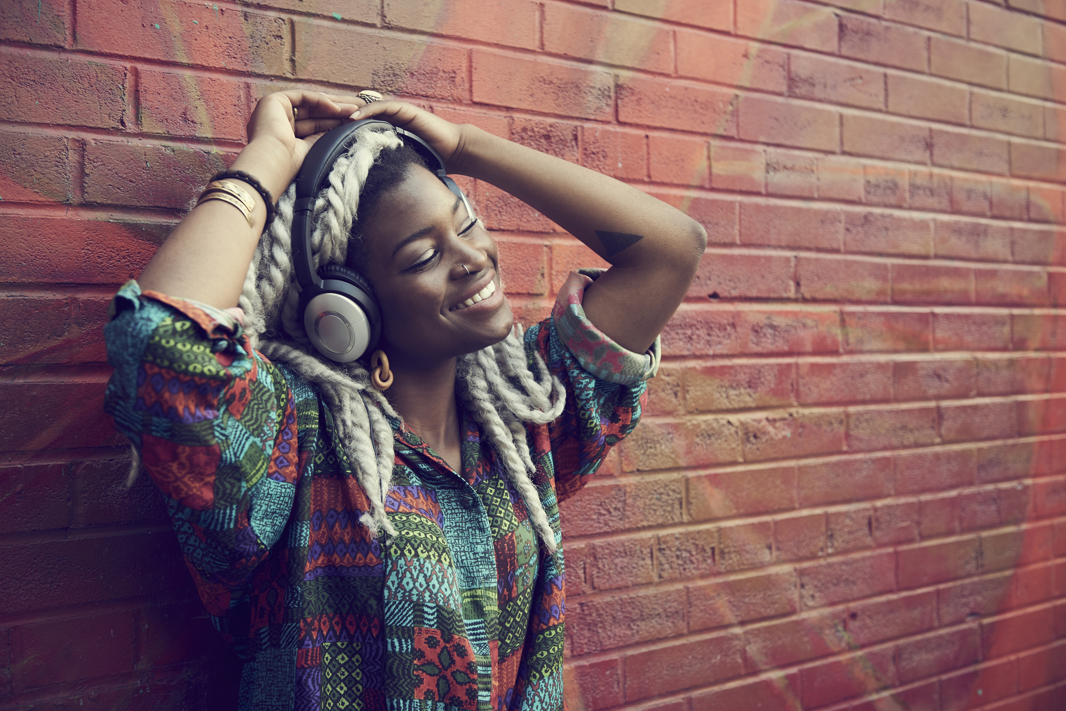 Black woman leaning on brick wall listening to headphones