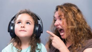 Angry Mother Shouting While Cute Girl Listening To Headphones Against Gray Background