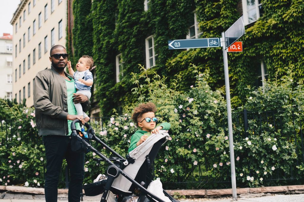 Father walking with children while pushing baby stroller against plants in city