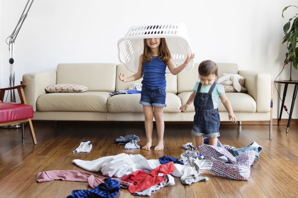 Young boy and girl with laundry basket over her head standing in front of sofa, laundry scattered on hardwood floor.