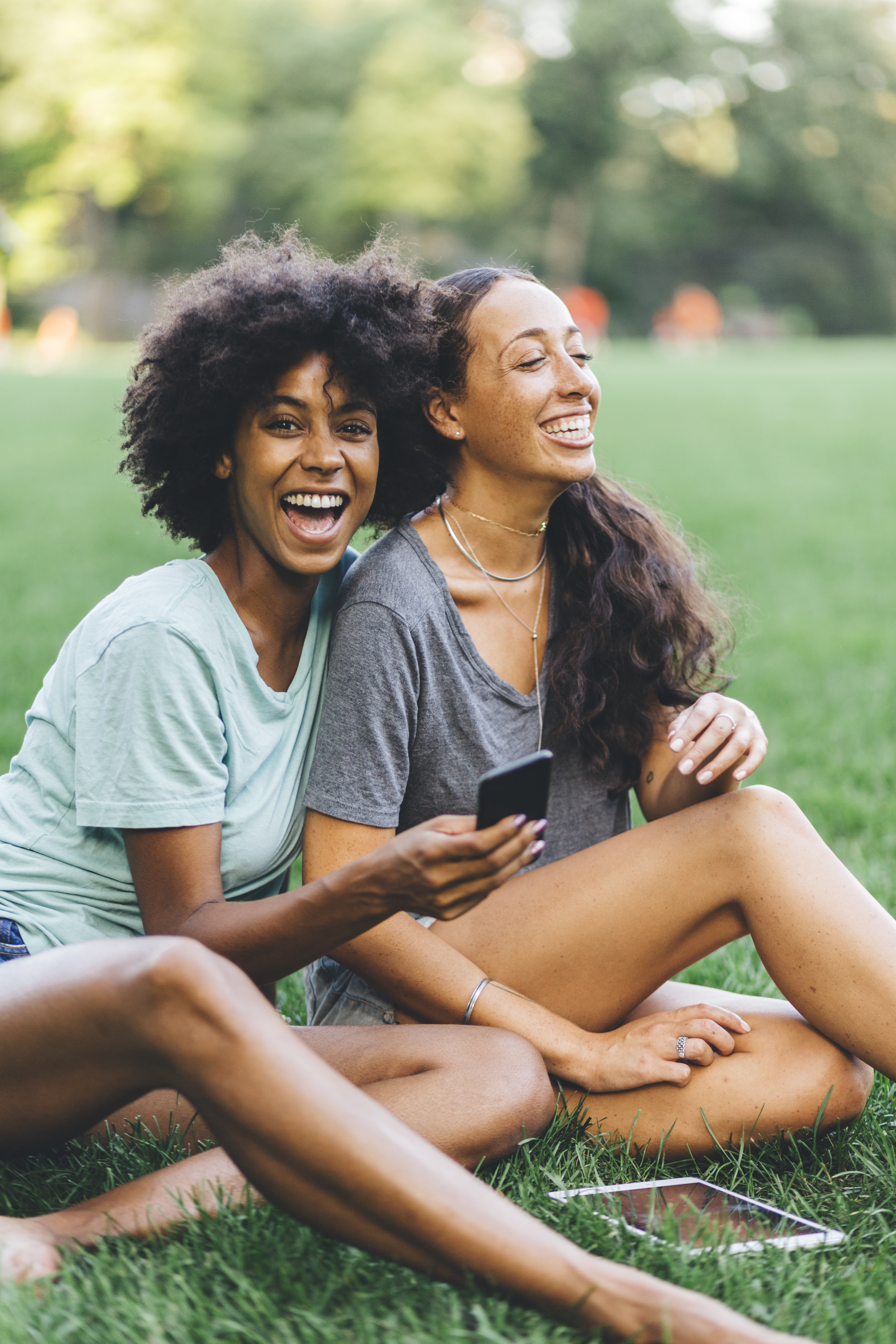 Two best friends sitting on a meadow of a park having fun with mini tablet