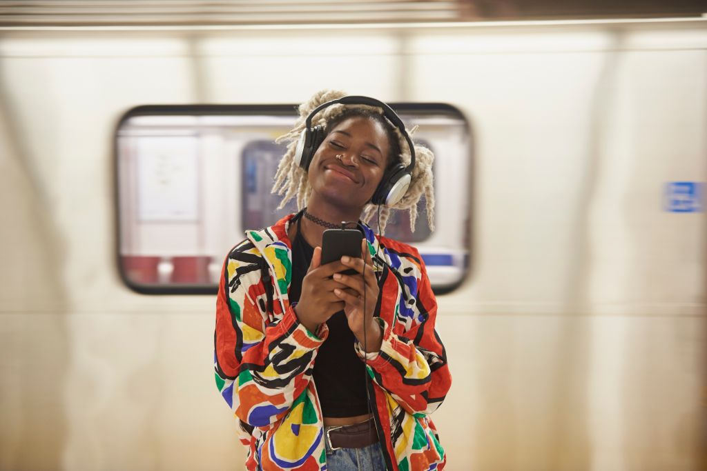 Black woman listening to cell phone with headphones near subway