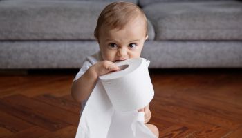 Baby boy with blond hair sitting on hardwood floor, playing with toilet paper rolls.
