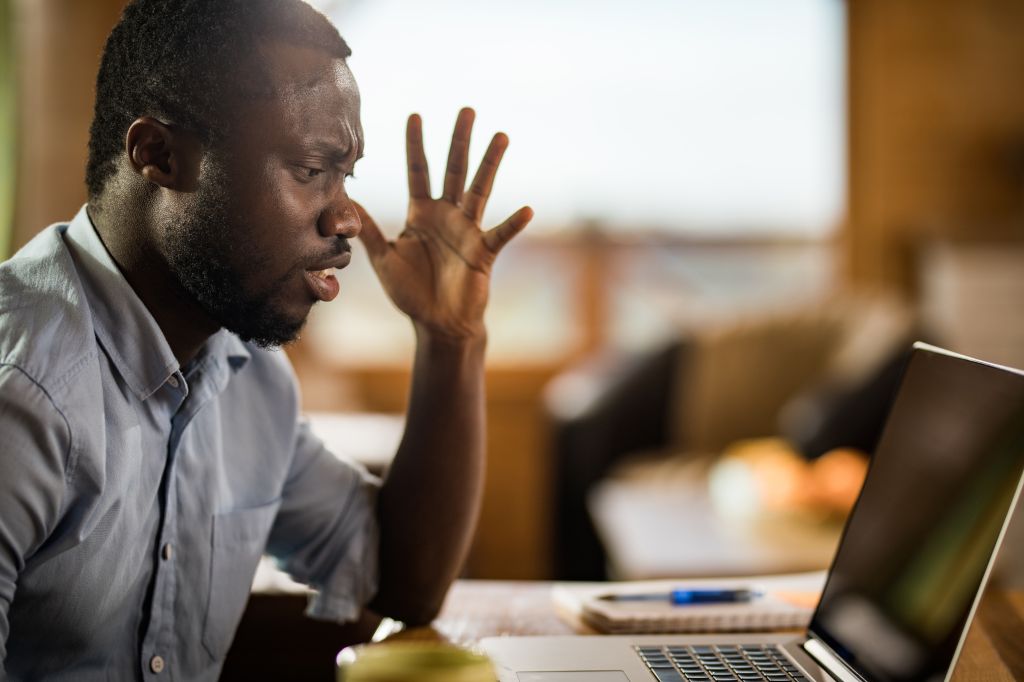 Frustrated black man reading annoying e-mail on laptop at home.