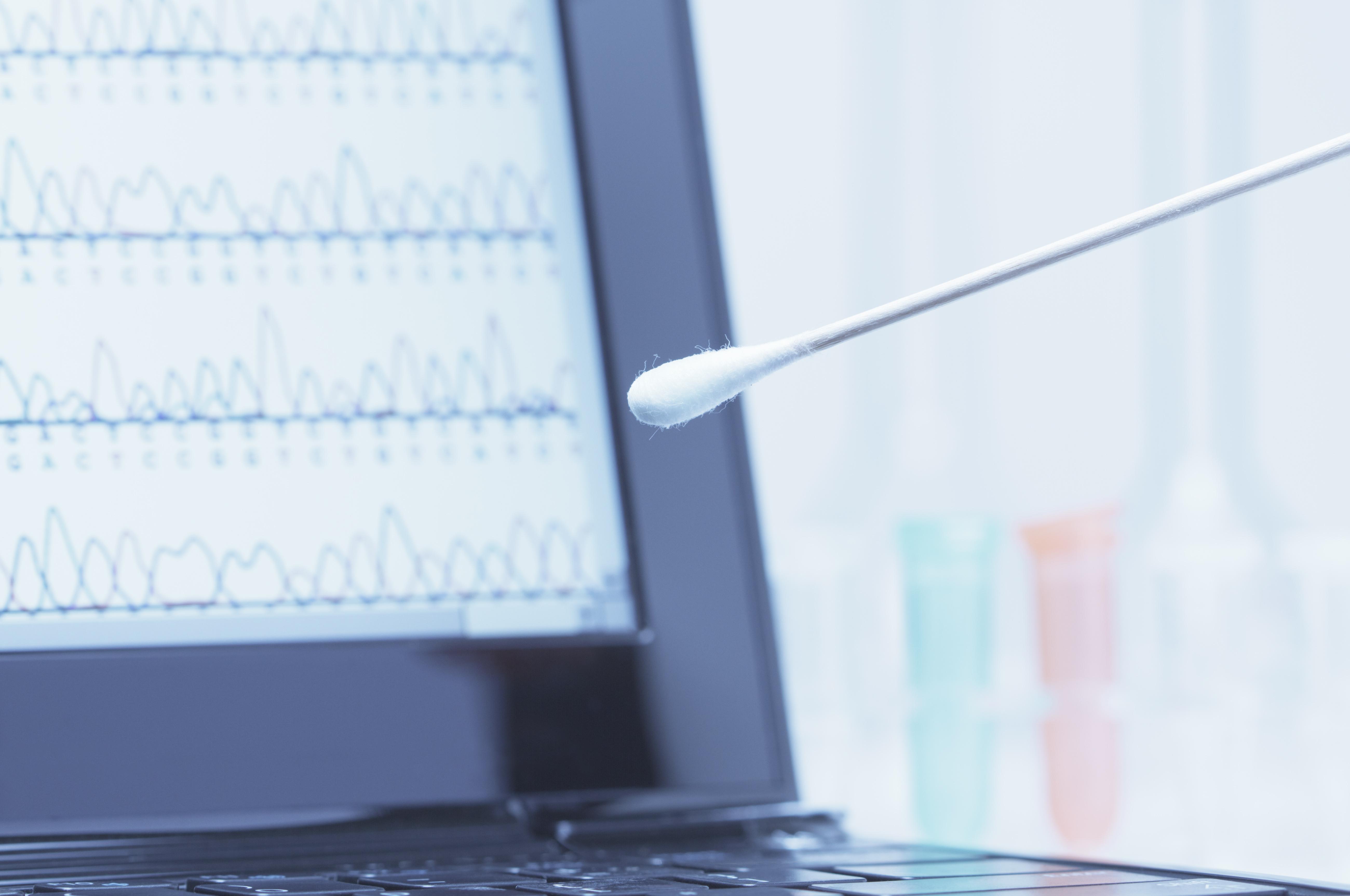 Cotton swab with saliva in front of a laptop screen that displays results of automated DNA sequencing