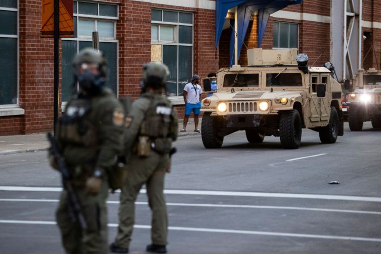 National Guard vehicles drive into downtown Louisville behind police officers in riot gear.