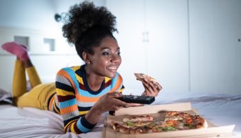 Afro woman spending lonely evening in bed with pizza and watching tv