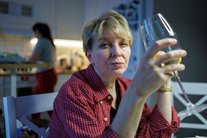 Mature blond woman wearing a red shirt is drinking white wine when her adult daughter is looking in a kitchen, blurred in the backdrop.