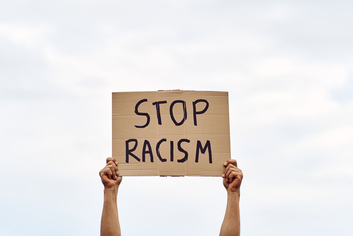 Unrecognizable man's hands holding a protest banner with the message STOP RACISM, with the sky in the background.