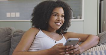 Beautiful young black woman using her phone while sitting on the couch in her living room at home. Happy female with afro thinking what to reply while texting or browsing through social media posts