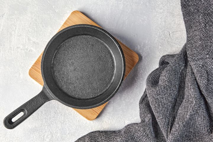Black cast iron empty frying pan for one person on a wooden stand and a gray kitchen towel on a gray concrete background