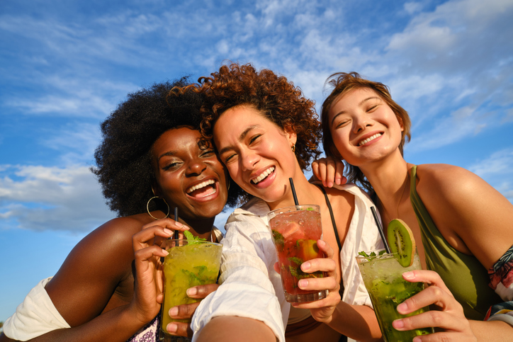 Three friends smiling at camera and drinking cocktails outdoors