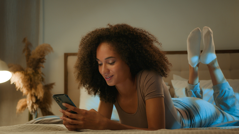 African American ethnic woman 20s girl curly hair scrolling mobile phone relaxing at home evening night insomnia smiling dreaming female lying on bed with smartphone gadget chatting online dating app