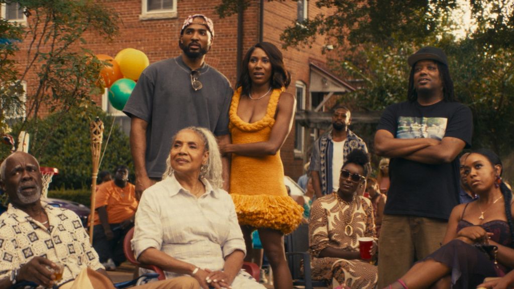 A group of people gathered outdoors, including an older woman in a white shirt, a woman in a yellow dress, and several men in casual clothing. Balloons and trees are visible in the background.