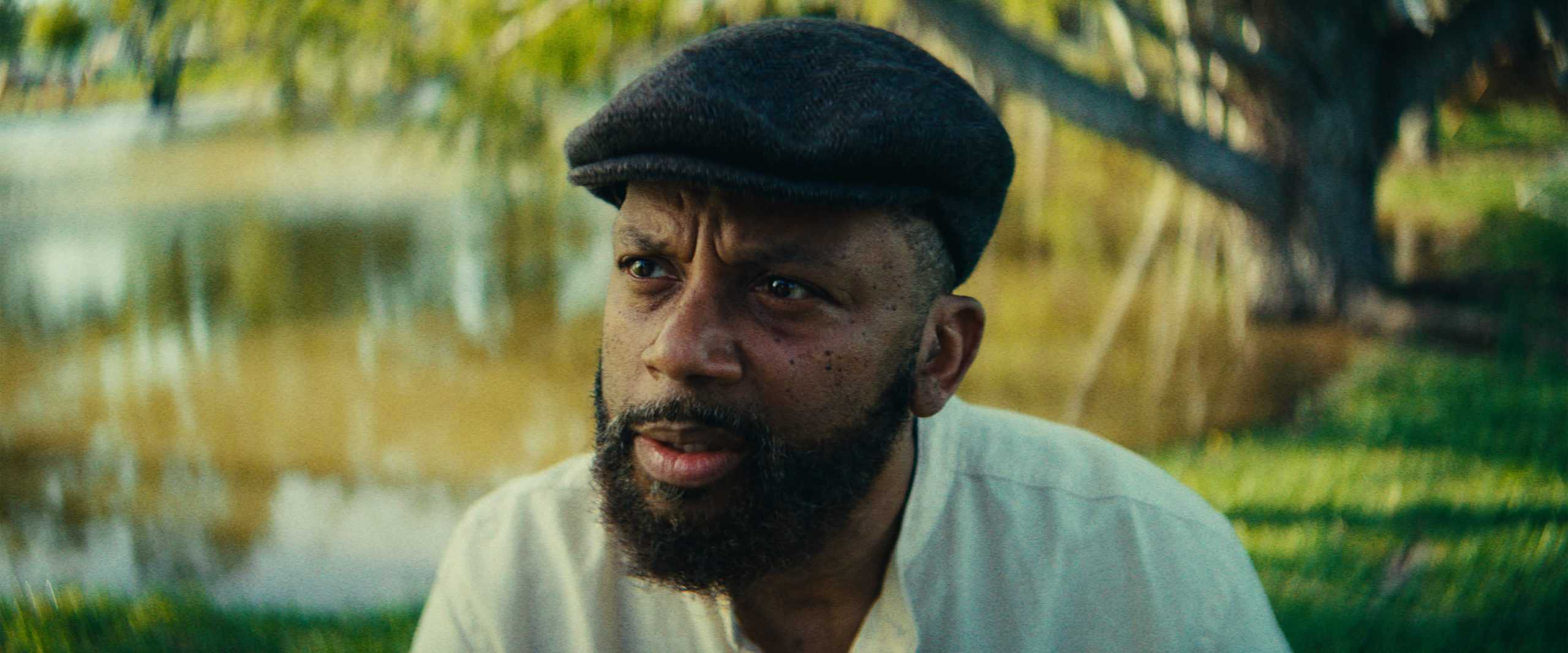 A man with a beard and hat looking directly at the camera in a natural outdoor setting.