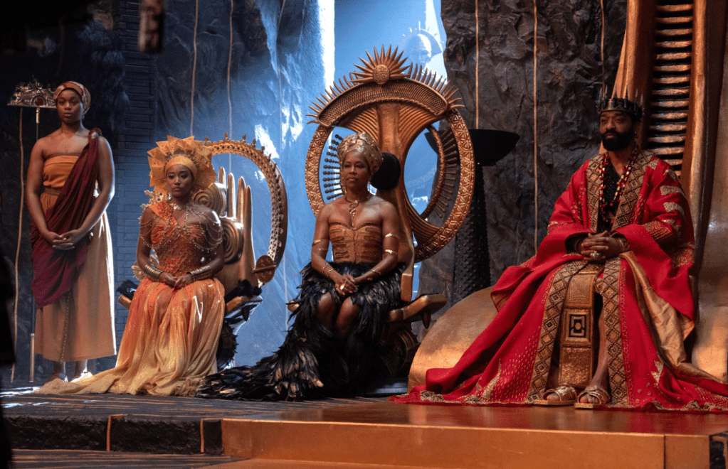 Ornate stage with elaborately dressed performers in traditional costumes, including feathered headdresses and flowing robes, against a backdrop of dramatic scenery.