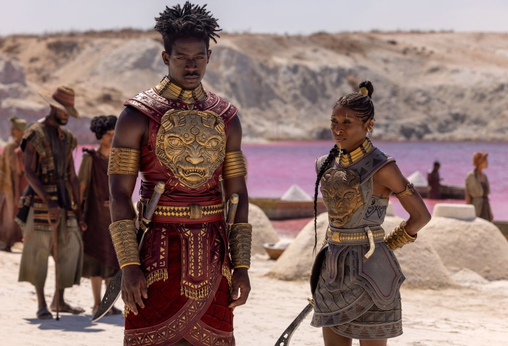Two people in elaborate tribal costumes stand in a desert landscape with a pink-colored lake in the background.