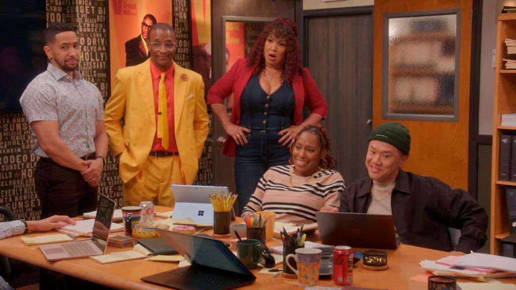 A group of five people in an office setting, including a man in a yellow suit, a woman in a red jacket, and others working at desks with laptops and office supplies.