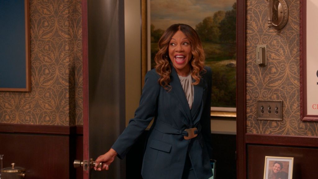 A smiling woman with curly brown hair wearing a navy blue blazer stands in a doorway against a backdrop of a landscape painting.