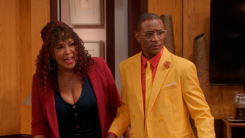 Two people, a woman in a red jacket and a man in a yellow suit, standing together and smiling in a room with wooden walls.