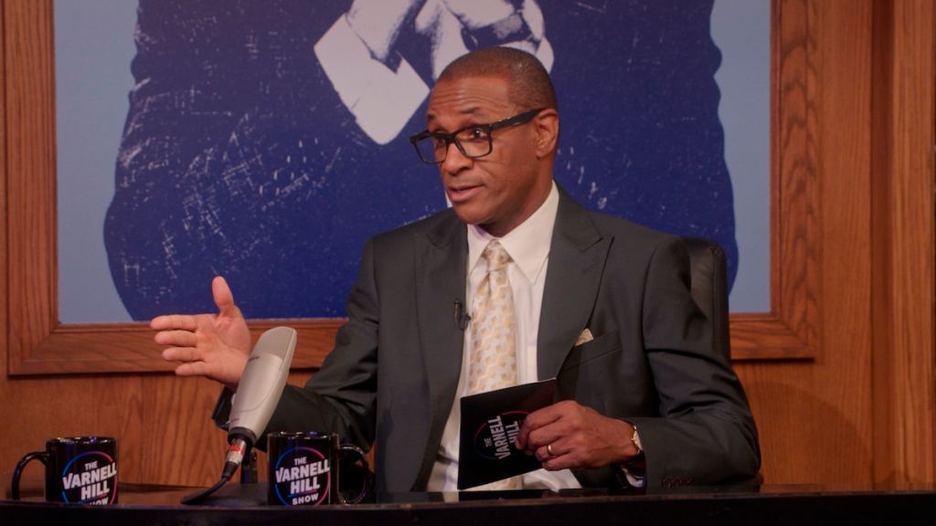A man in a suit and glasses stands at a podium with "Varnell Hill Show" branding, gesturing as he speaks.