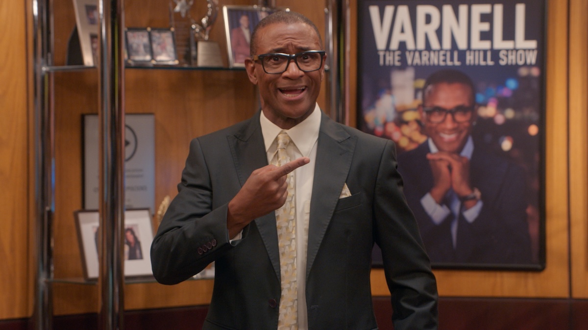 A smiling man in a suit and glasses stands in front of a poster for "The Varnell Hill Show" on Paramount+.