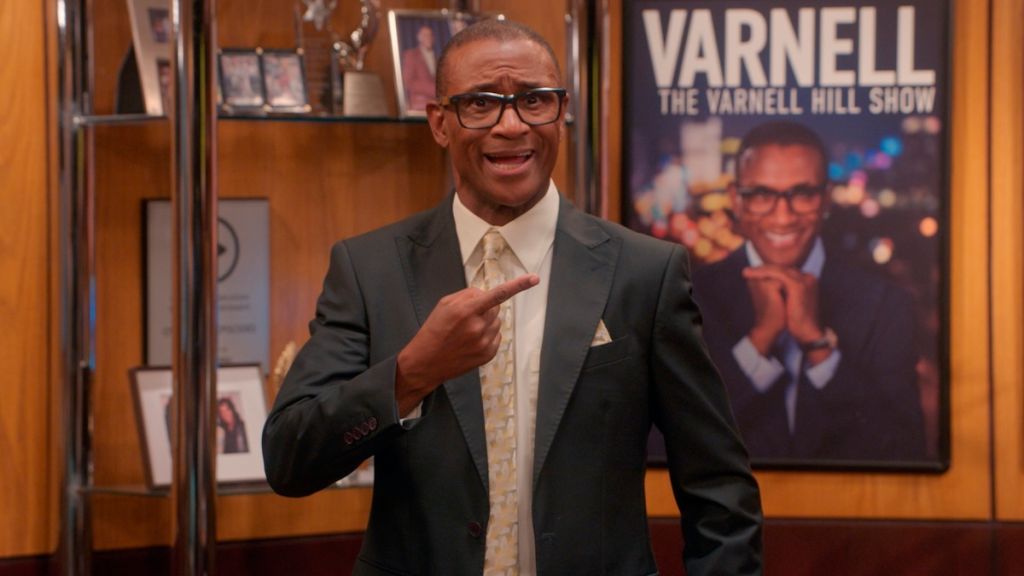 A smiling man in a suit and glasses stands in front of a poster for "The Varnell Hill Show" on Paramount+.