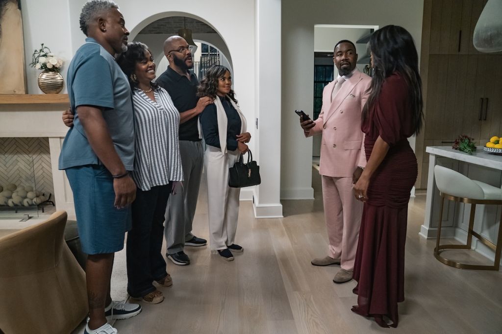 A group of six adults, including two men and four women, standing together in a home interior. The individuals are dressed in formal attire, with one man wearing a pink suit.