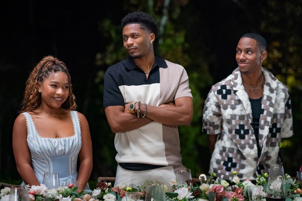 Three young adults, two men and one woman, standing together outdoors near a table with flowers and drinks.