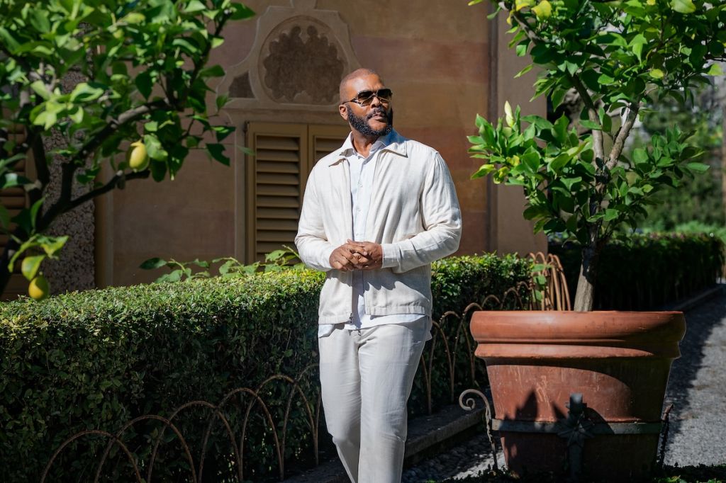 A man with a beard wearing a white outfit stands in a garden with lush greenery and a decorative architectural feature in the background.