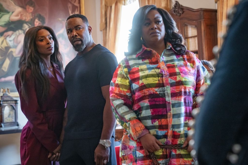 Three people standing in a room, two women and one man. The woman on the left is wearing a burgundy dress, the man in the middle is wearing a black shirt, and the woman on the right is wearing a colorful plaid shirt.