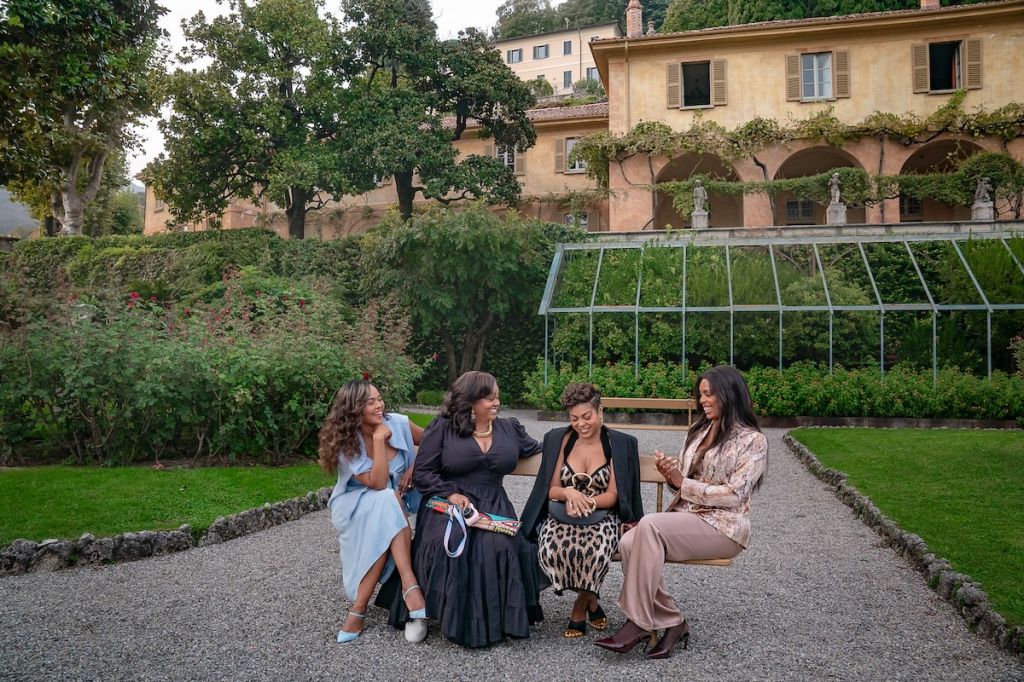 Four women in formal attire sitting in a lush garden with a historic building in the background.