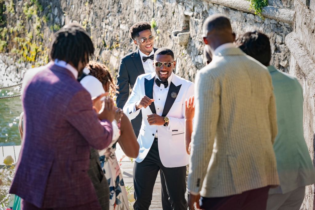 A group of well-dressed men, some in tuxedos, laughing and conversing outdoors in a scenic setting with a stone wall in the background.