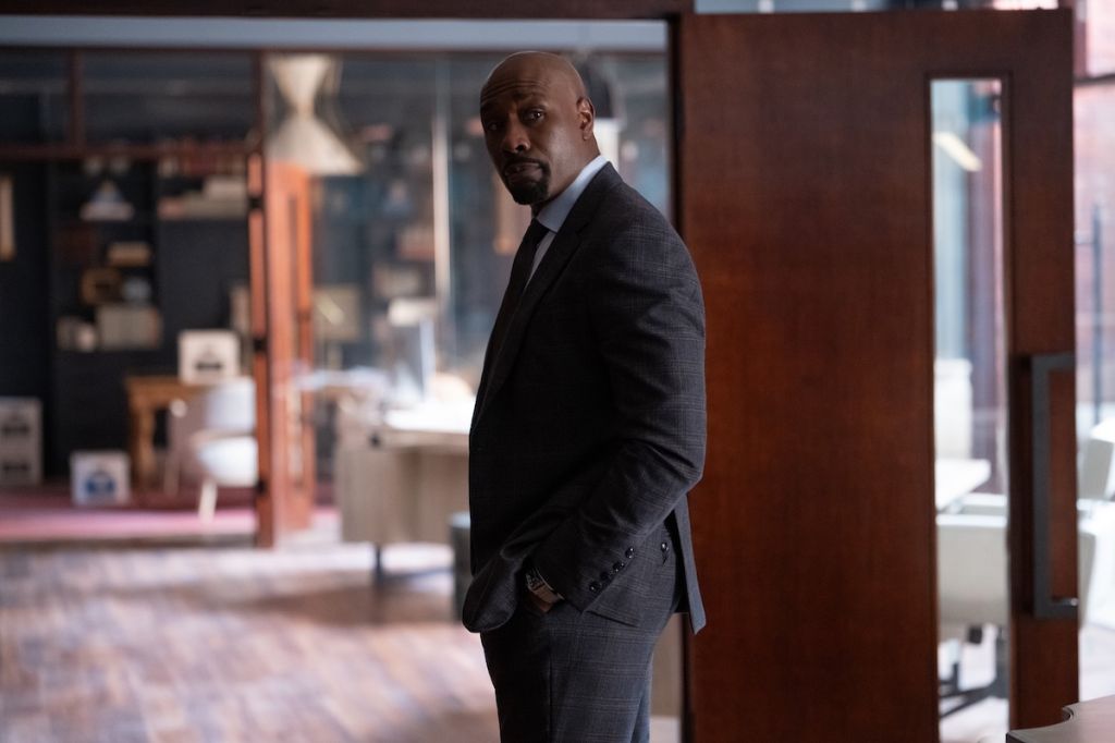 A well-dressed Black man in a suit standing in a room with glass walls and furniture.