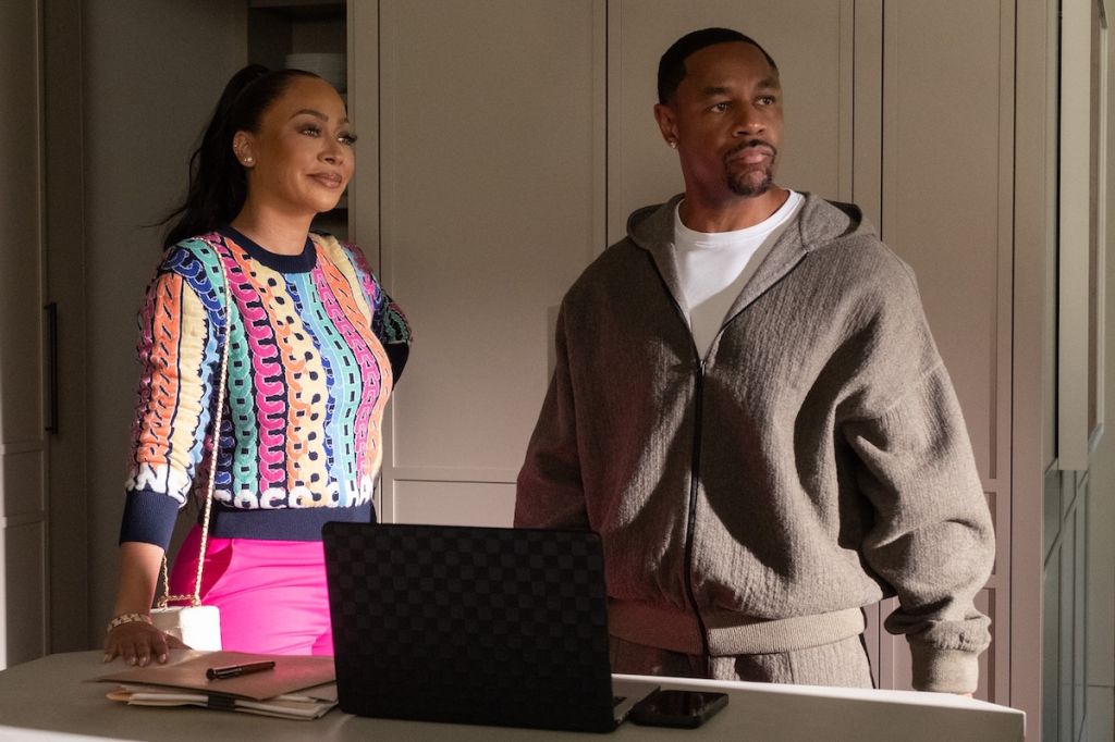 Two people, a woman wearing a colorful patterned top and a man wearing a gray hooded sweater, standing in front of a laptop on a table.