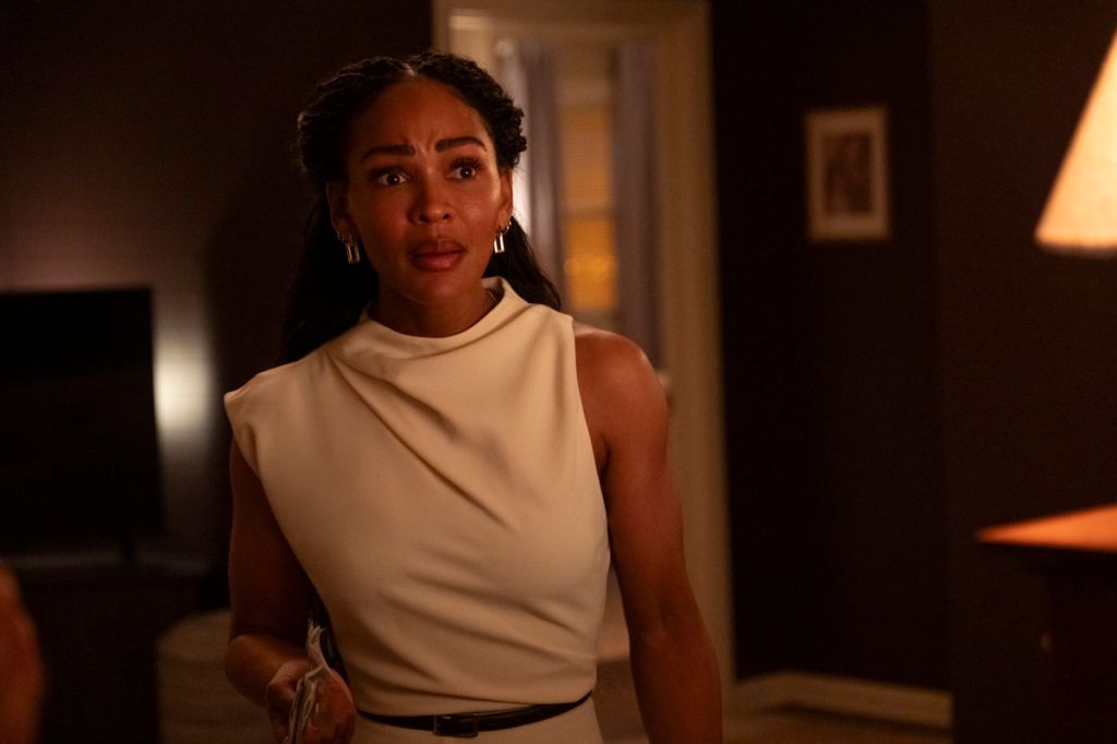 A woman with curly hair wearing a beige sleeveless top stands in a dimly lit room, looking concerned.