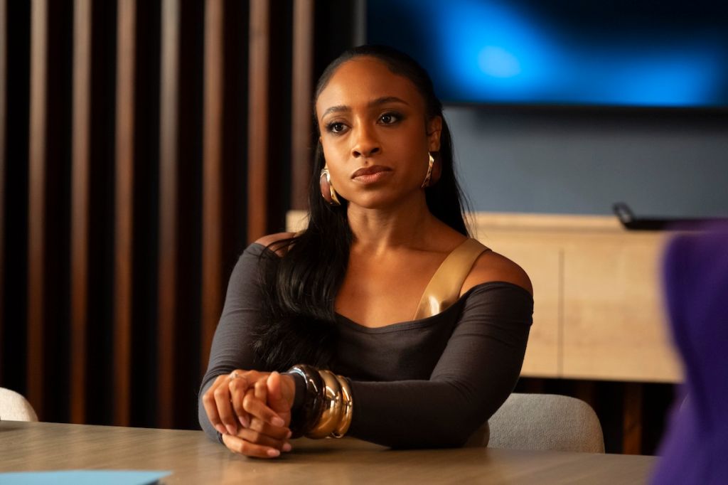 A woman with long dark hair wearing a black off-the-shoulder top sits at a table with a serious expression.